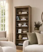 Taylor Oak Bookcase in natural oak with dark metal supports and open shelving, styled with books, ceramics and decorative objects in a neutral living room.